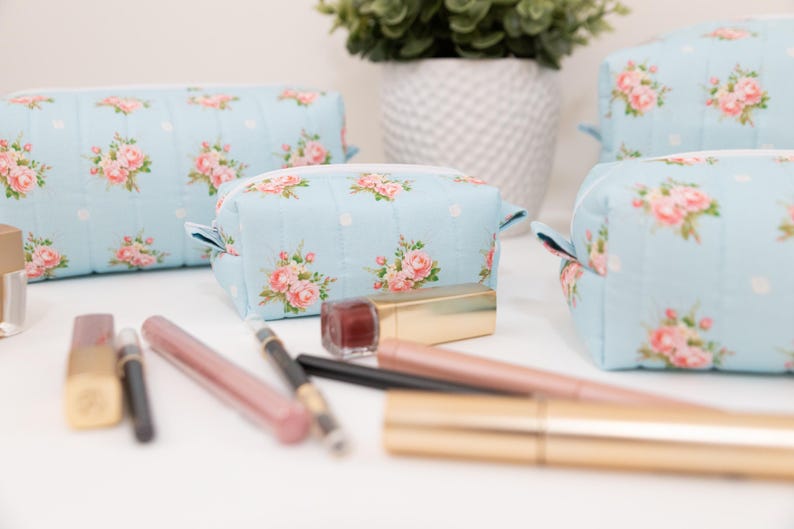 May include: Three light blue quilted cosmetic bags featuring a repeating pattern of pink roses and white polka dots, each with a white zipper closure. Several makeup items, including lipsticks and eyeliner pencils, are arranged in the foreground.