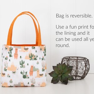 May include: A small, reversible tote bag with orange handles. The bag is made of white fabric with a pattern of llamas, cacti, and mountains. The text "Bag is reversible. Use a fun print for the lining and it can be used all year round." is visible in the image.
