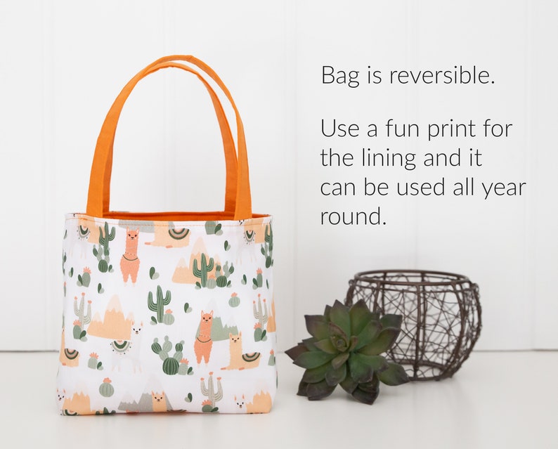May include: A small, reversible tote bag with orange handles. The bag is made of white fabric with a pattern of llamas, cacti, and mountains. The text "Bag is reversible. Use a fun print for the lining and it can be used all year round." is visible in the image.
