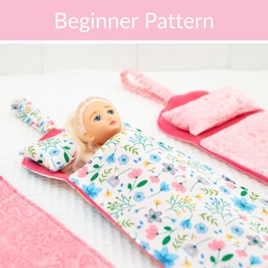 May include: A doll lying in a fabric sleeping bag with a white base and a floral pattern of blue, pink, and green flowers. The set includes a matching pillow and a fabric loop for hanging. The text "Beginner Pattern" is visible at the top.