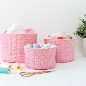 May include: Three pink fabric storage baskets of varying sizes, filled with yarn, fabric scraps, and sewing notions. A pair of gold scissors and spools of thread are in the foreground. A potted plant is in the background.