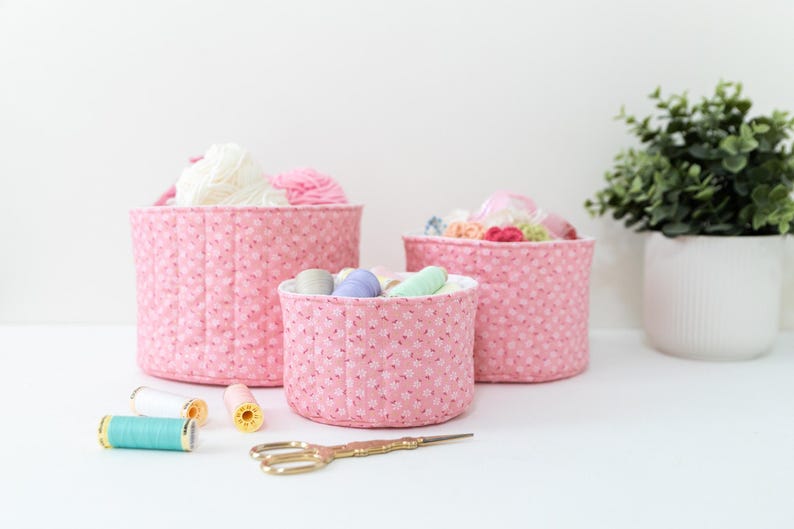May include: Three pink fabric storage baskets of varying sizes, filled with yarn, fabric scraps, and sewing notions. A pair of gold scissors and spools of thread are in the foreground. A potted plant is in the background.