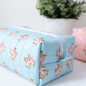 May include: A rectangular quilted pouch in light blue fabric featuring a repeating pattern of pink roses and small white polka dots, finished with a white zipper closure. A second identical pouch in pink is partially visible in the background.