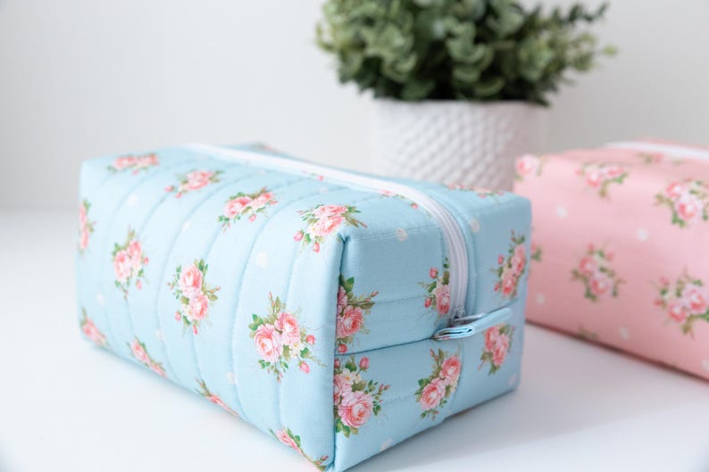 May include: A rectangular quilted pouch in light blue fabric featuring a repeating pattern of pink roses and small white polka dots, finished with a white zipper closure. A second identical pouch in pink is partially visible in the background.