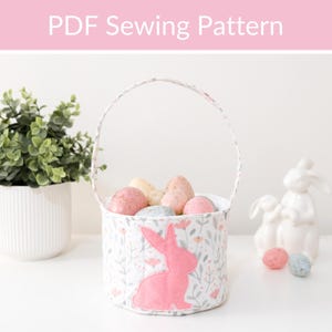 May include: A white Easter basket with a pink bunny applique and floral pattern, filled with decorated eggs. The basket has a handle and is next to a white ceramic bunny figurine and a potted plant. Text at the top says "PDF Sewing Pattern."