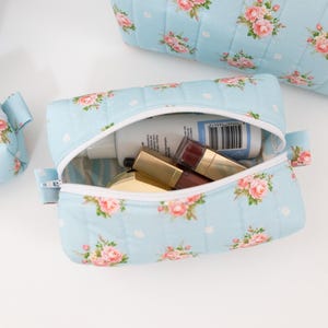 May include: A light blue quilted cosmetic bag featuring a pink rose floral pattern and white polka dots. The bag has a white zipper closure and contains various small personal care items, including a bottle with a visible barcode label.