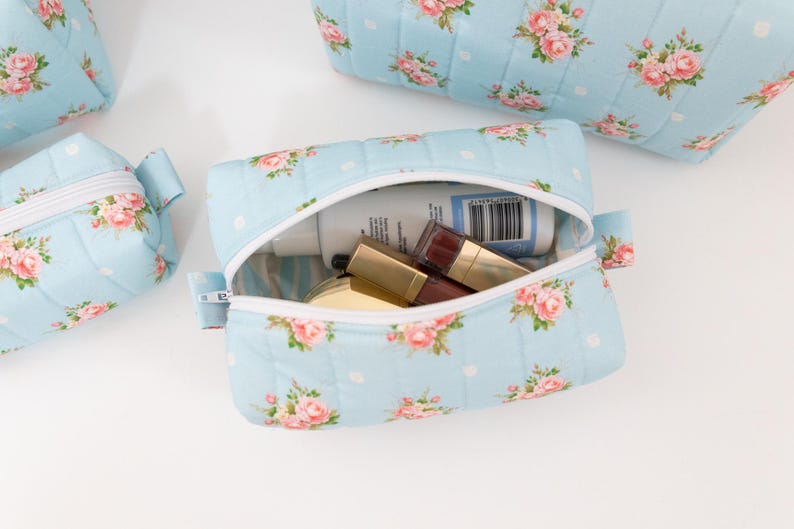 May include: A light blue quilted cosmetic bag featuring a pink rose floral pattern and white polka dots. The bag has a white zipper closure and contains various small personal care items, including a bottle with a visible barcode label.