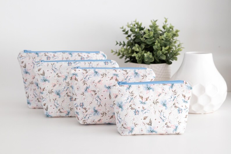 Four beginner zip pouches in soft blue and pink floral fabric, arranged in graduated sizes from large to small on a white table with a plant and white pot.