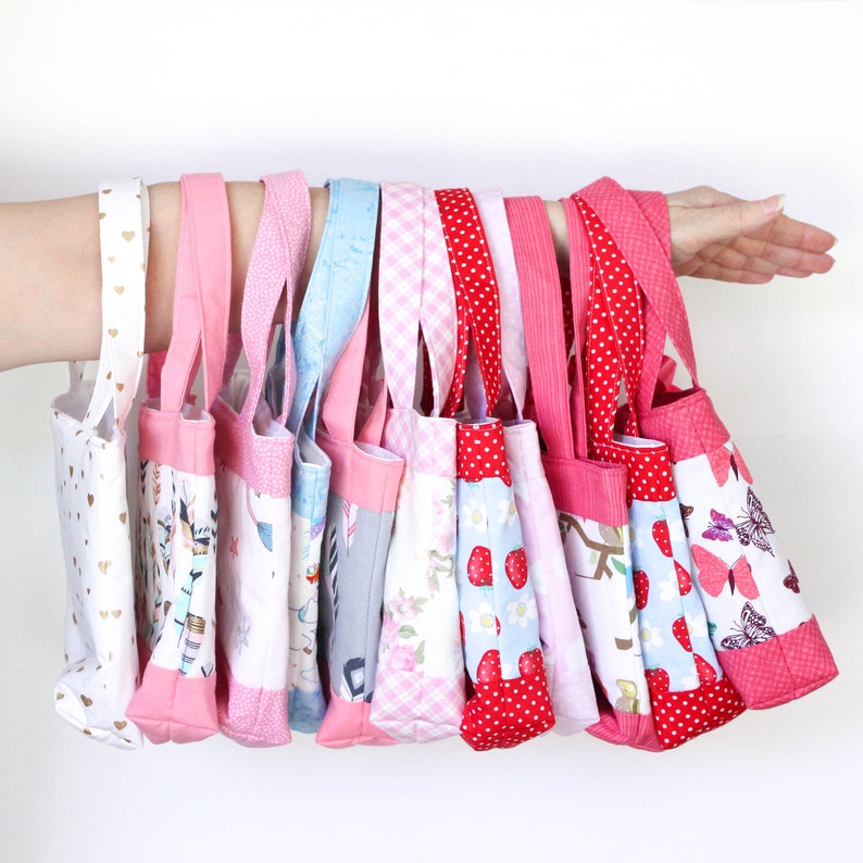 May include: A collection of 10 small pink and red fabric tote bags with various patterns, including hearts, flowers, and butterflies. The bags are lined up in a row, hanging from a hand.