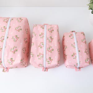 May include: Four rectangular zippered pouches in descending sizes, featuring a light pink fabric with a repeating pattern of small pink roses, green leaves, and white polka dots. Each pouch has a white zipper closure running down the center.