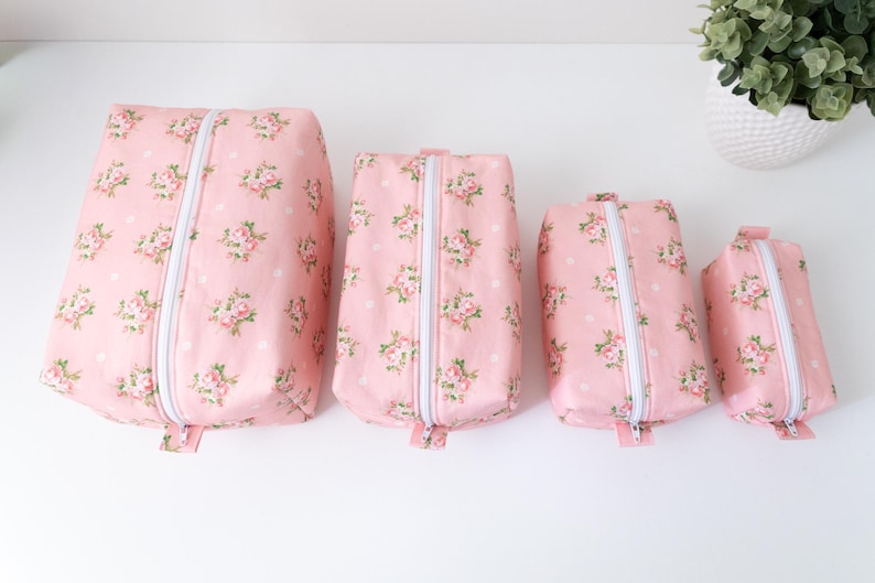 May include: Four rectangular zippered pouches in descending sizes, featuring a light pink fabric with a repeating pattern of small pink roses, green leaves, and white polka dots. Each pouch has a white zipper closure running down the center.