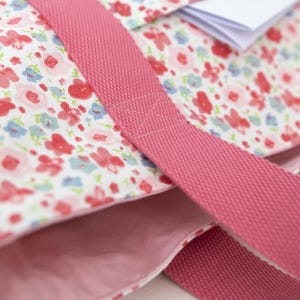 Puede incluir: Un bolso de tela con base blanca y un patr&oacute;n repetitivo de peque&ntilde;as flores rojas, rosas y azul claro. El bolso incluye asas anchas de tela tejida en color rosa s&oacute;lido y un forro interior rosa s&oacute;lido, con una peque&ntilde;a etiqueta blanca visible.