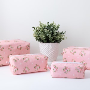 May include: A set of four rectangular zippered pouches in varying sizes, all featuring a light pink fabric base with a repeating pattern of small floral bouquets and white polka dots, each equipped with a white zipper closure along the top edge.