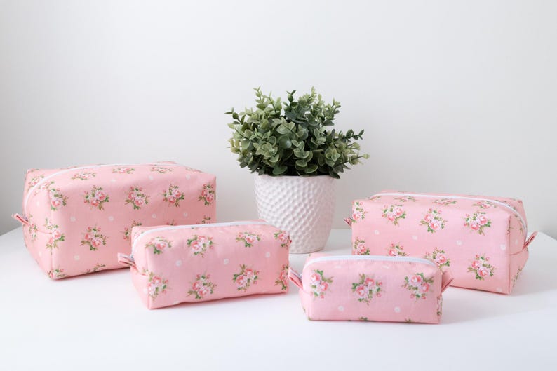 May include: A set of four rectangular zippered pouches in varying sizes, all featuring a light pink fabric base with a repeating pattern of small floral bouquets and white polka dots, each equipped with a white zipper closure along the top edge.