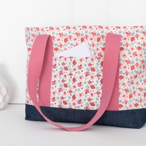 Puede incluir: Un bolso tote con un estampado floral de flores rojas, rosas y azules sobre fondo blanco. El bolso tiene una base de tela vaquera azul oscuro y dos asas de tela rosa s&oacute;lido. Una tarjeta rectangular blanca est&aacute; insertada en el bolsillo frontal.