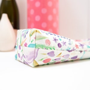May include: A rectangular pencil case with a floral pattern in watercolor style. The design features purple, pink, and teal flowers and leaves on a white background. The case is made of fabric.