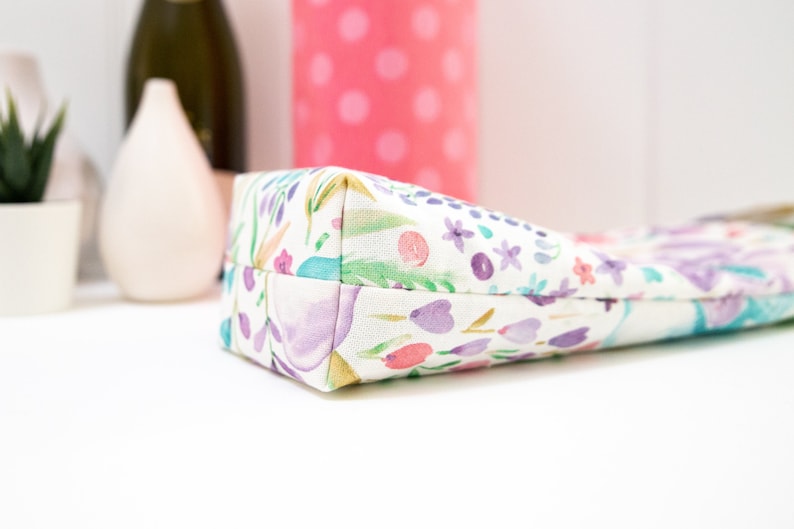 May include: A rectangular pencil case with a floral pattern in watercolor style. The design features purple, pink, and teal flowers and leaves on a white background. The case is made of fabric.