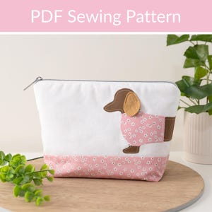 May include: A white zippered pouch with a pink and white floral print on the bottom. A brown dachshund with a pink floral print shirt is sewn onto the front of the pouch. The pouch is sitting on a wooden surface.