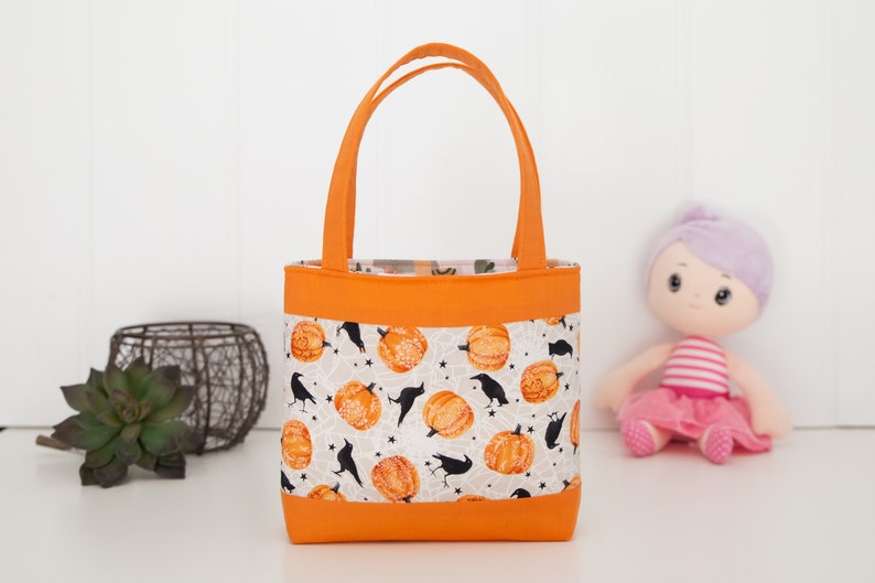 May include: A small orange tote bag with a white fabric lining featuring a pattern of black crows and orange pumpkins. The bag has two orange handles.