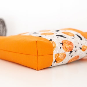 May include: Orange tote bag with a white fabric lining featuring a pattern of orange pumpkins, black bats, and white stars on a gray background.