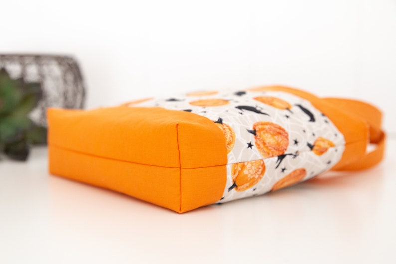 May include: Orange tote bag with a white fabric lining featuring a pattern of orange pumpkins, black bats, and white stars on a gray background.