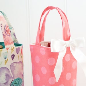 May include: Two fabric wine bottle bags. One is pink with white polka dots and a white bow. The other has a floral pattern with purple, pink, and blue flowers. Both have handles for carrying.