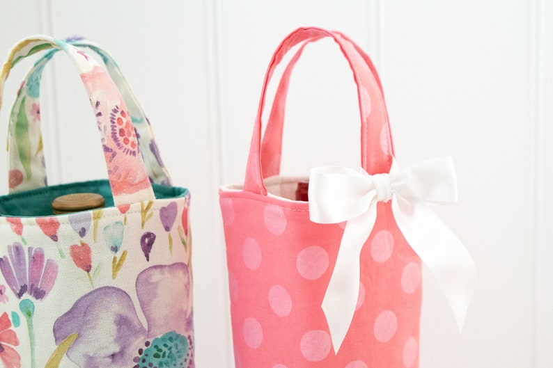 May include: Two fabric wine bottle bags. One is pink with white polka dots and a white bow. The other has a floral pattern with purple, pink, and blue flowers. Both have handles for carrying.