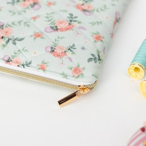 K&ouml;nnte beinhalten: Eine gesteppte Tasche mit floralem Muster und goldenem Rei&szlig;verschluss, mit rosa und pfirsichfarbenen Rosen auf hellgr&uuml;nem Hintergrund. Zwei Garnrollen, eine hellblaue und eine rosa, liegen neben der Tasche.