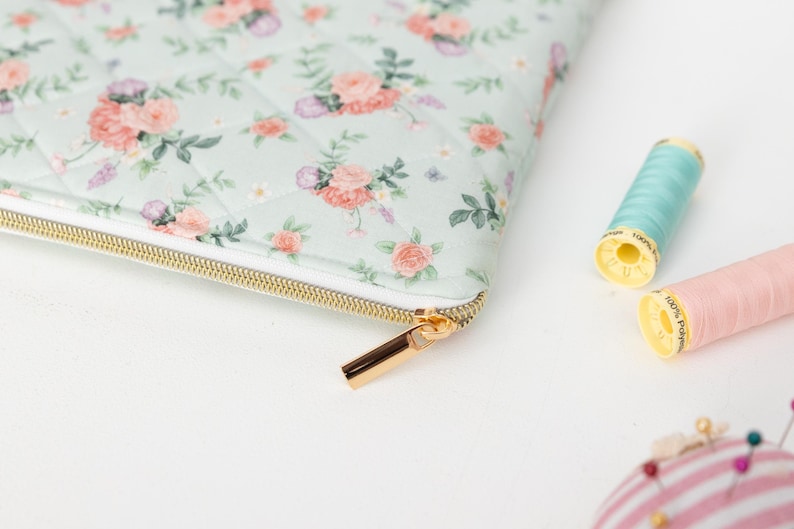 K&ouml;nnte beinhalten: Eine gesteppte Tasche mit floralem Muster und goldenem Rei&szlig;verschluss, mit rosa und pfirsichfarbenen Rosen auf hellgr&uuml;nem Hintergrund. Zwei Garnrollen, eine hellblaue und eine rosa, liegen neben der Tasche.