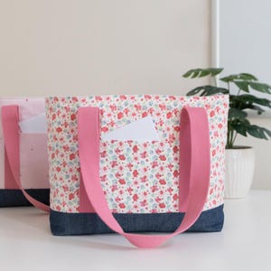 Puede incluir: Dos bolsos tote con asas de tela rosa y base de mezclilla azul oscuro. El bolso delantero presenta una tela blanca con un estampado floral rojo y azul y un bolsillo peque&ntilde;o. El bolso trasero es rosa claro con un bolsillo a juego.