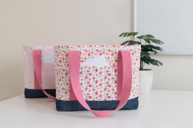 Puede incluir: Dos bolsos tote con asas de tela rosa y base de mezclilla azul oscuro. El bolso delantero presenta una tela blanca con un estampado floral rojo y azul y un bolsillo peque&ntilde;o. El bolso trasero es rosa claro con un bolsillo a juego.