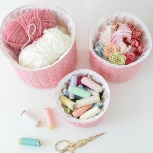 May include: Three pink fabric baskets filled with craft supplies. The largest basket holds pink and white yarn. The second basket contains fabric flowers and bows. The smallest basket is filled with spools of thread. Gold scissors and thread spools are also visible.