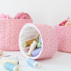 May include: Three pink fabric storage containers, one round and two rectangular, are filled with yarn and spools of thread. The containers have a floral pattern. Spools of thread in various colors and a pair of gold scissors are scattered around.