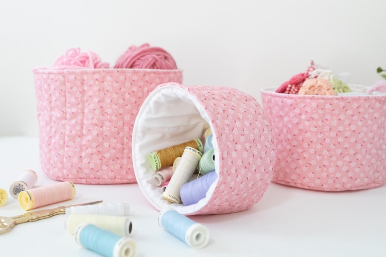 May include: Three pink fabric storage containers, one round and two rectangular, are filled with yarn and spools of thread. The containers have a floral pattern. Spools of thread in various colors and a pair of gold scissors are scattered around.