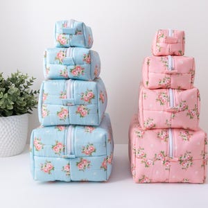 May include: Two stacks of four rectangular zippered pouches, one stack in light blue and one in light pink. Each pouch features a repeating floral pattern of pink roses with green leaves and small white dots. Each pouch has a white zipper closure.