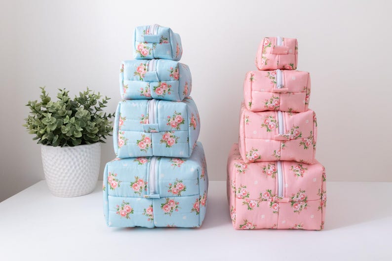 May include: Two stacks of four rectangular zippered pouches, one stack in light blue and one in light pink. Each pouch features a repeating floral pattern of pink roses with green leaves and small white dots. Each pouch has a white zipper closure.