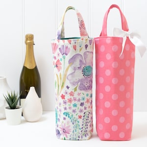 May include: Two wine bottle bags. One is pink with white polka dots and a white bow. The other is floral with purple, pink, and blue flowers. Both have handles and are designed to hold a wine bottle.