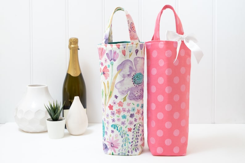 May include: Two wine bottle bags. One is pink with white polka dots and a white bow. The other is floral with purple, pink, and blue flowers. Both have handles and are designed to hold a wine bottle.