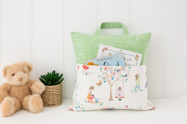 Puede incluir: Una almohada blanca con un asa verde y un estampado colorido de ni&ntilde;os jugando en un jard&iacute;n. La almohada tiene un bolsillo en la parte delantera que contiene dos libros, uno titulado "Mr. Bear Says Goodnight" y el otro titulado "The Tale of the Unlucky Unicorn".