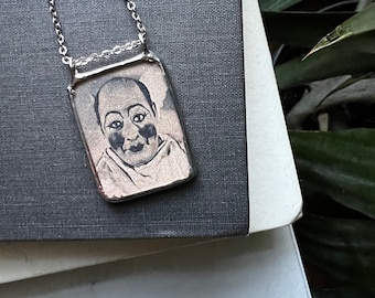 May include: Silver-toned pendant necklace featuring a rectangular pendant with a portrait of a person. The portrait is in black and white, with a smiling face. The necklace chain is also silver.
