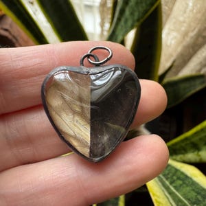 May include: Heart-shaped pendant with a split design. One half features a light brown, textured pattern, while the other is a dark, glossy black. The pendant has a silver-colored metal frame and a small loop at the top for a chain.