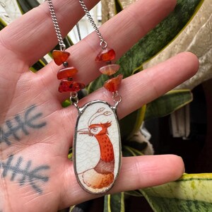 May include: A silver chain necklace featuring an oval pendant with a bird illustration in shades of red and orange. The necklace includes orange-red gemstone accents. The pendant is set against a white background with a crackle effect.