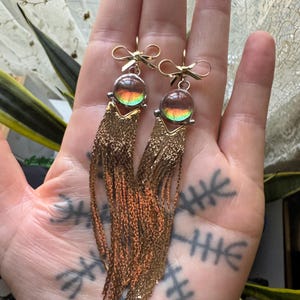 May include: A pair of gold-toned earrings with a bow design at the top. Each earring features a round, iridescent bead and long, dangling chains. The chains are a mix of gold and copper colors, creating a unique, eye-catching accessory.