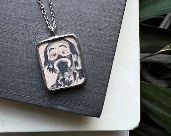 May include: A silver-toned pendant necklace featuring a rectangular charm with a black and white illustration of a person with a painted face. The pendant hangs on a delicate chain, resting on a dark gray surface.