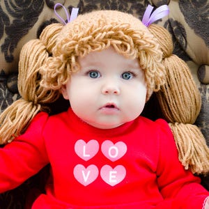 May include: A red long-sleeved shirt featuring four pink hearts arranged in a square, each containing a white letter to spell the word LOVE. The shirt is paired with a tan yarn wig styled with two pigtails and accented with small purple ribbons.