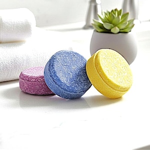 May include: Three round, solid shampoo bars in shades of purple, blue, and yellow are displayed on a white countertop. A white towel and a small succulent in a gray pot are also visible. The bars have a textured surface.