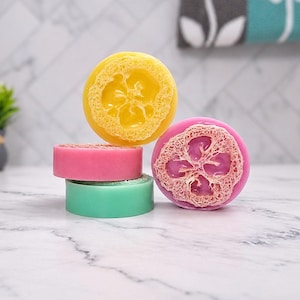May include: A collection of four round soap bars featuring embedded natural loofah sponges. The soaps are colored in bright yellow, pink, and light green. One pink soap is stacked beneath a yellow soap, while another pink soap stands upright.
