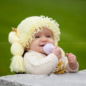 A toddler in a Halloween costume wearing a crocheted cabbage patch wig with yarn pigtails. The wig mimics the classic Cabbage Patch Kids doll hairstyle, with the pigtails tied off at the sides, giving the toddler a cute and nostalgic look.