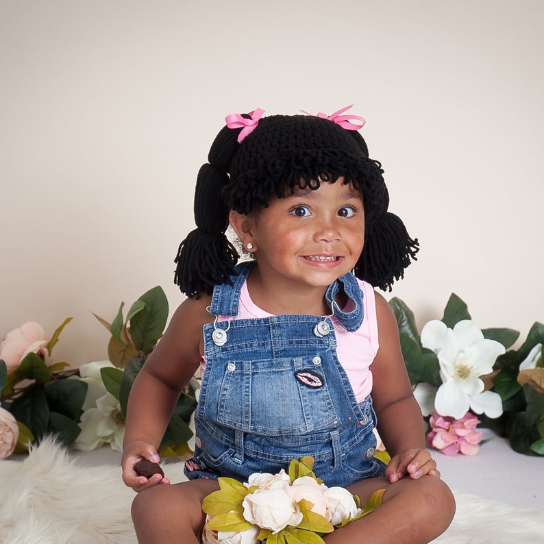 A baby wearing a handmade Halloween hat designed to look like a Cabbage Patch doll's hair. The crocheted hat mimics baby doll hair with yarn strands pulled neatly into pigtails, capturing the charming and playful essence of a Cabbage Patch Kids doll.