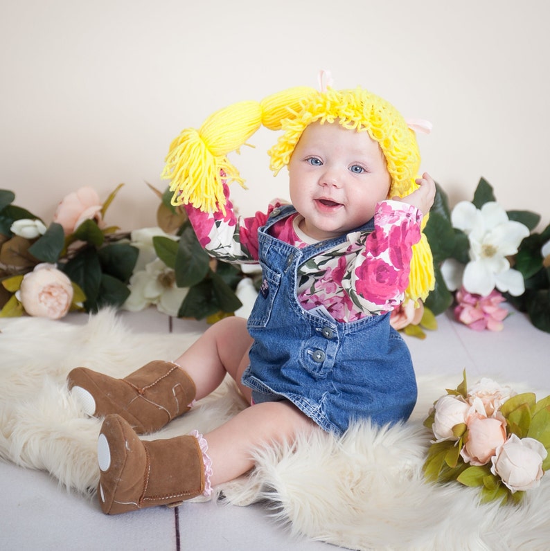 A baby dressed for Halloween, wearing a handmade crocheted cabbage patch hat with brown yarn pigtails. The hat resembles the iconic Cabbage Patch Kids doll hairstyle, adding a playful and adorable touch to the baby’s costume.